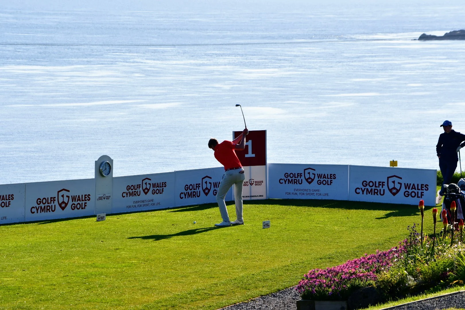First tee, blue sea in the background, surrounded by Wales Golf signage with a competitor playing his first shot.